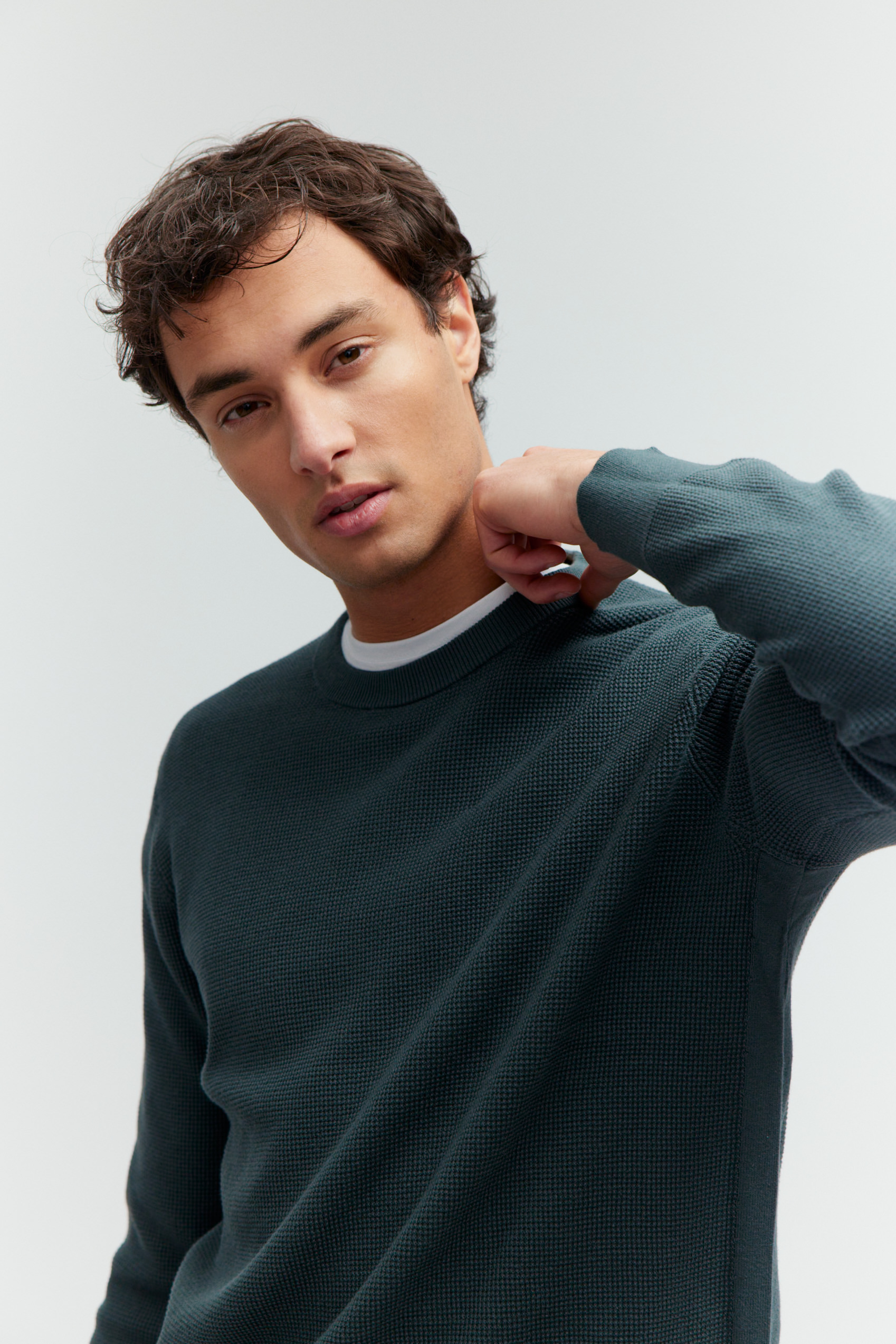 Porträt eines Models im dunkelgrünen Strukturpullover, die Hand am Kragen.