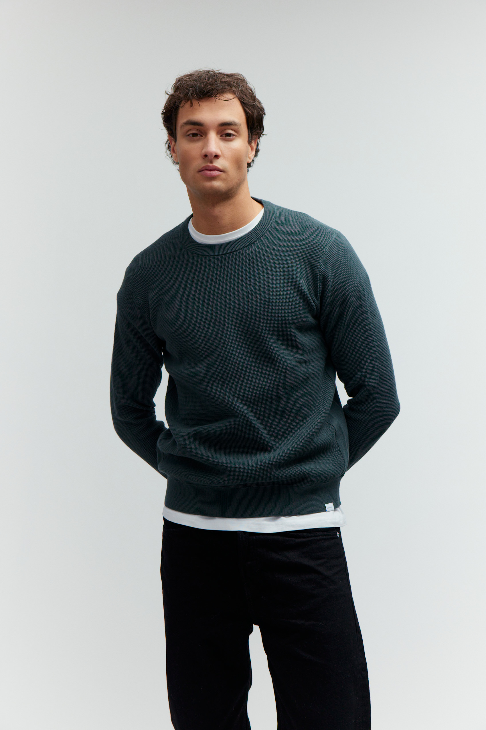 Model in dunkelgrünem Strukturpullover und schwarzer Jeans vor hellem Hintergrund.