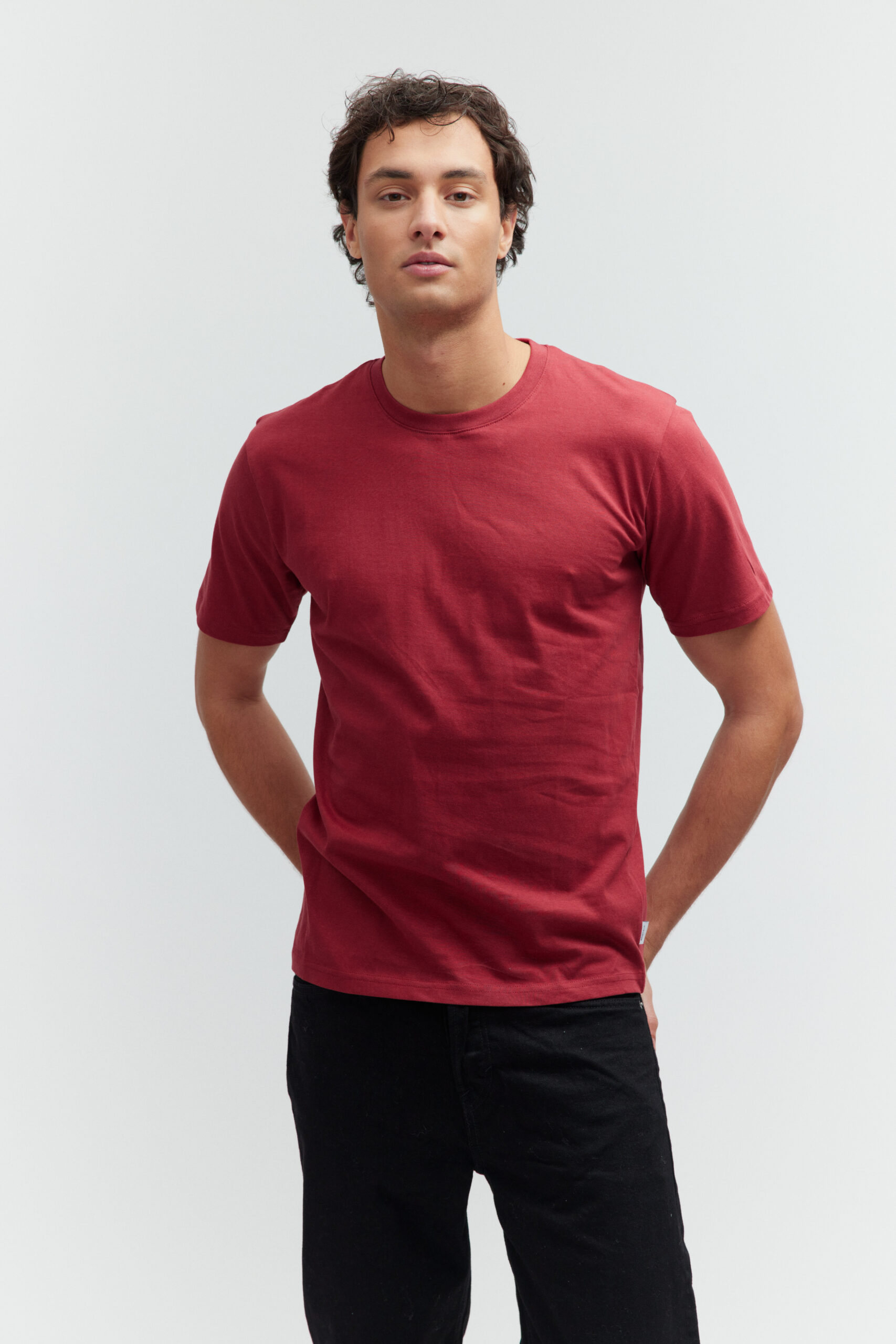 Model in bordeauxrotem T-Shirt und schwarzer Jeans vor hellem Hintergrund.