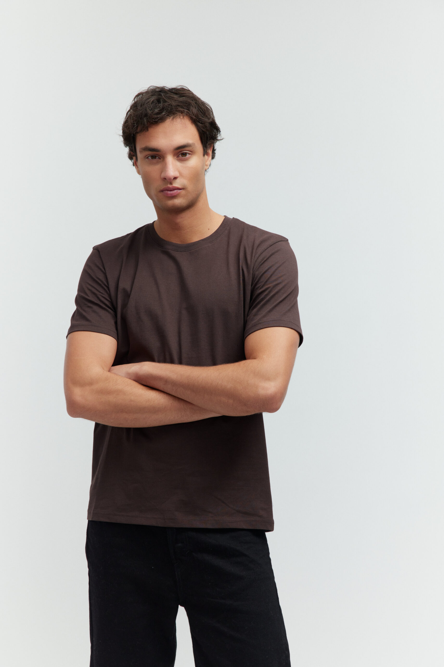 Model in moccabraunem T-Shirt und schwarzer Jeans, die Arme vor der Brust verschränkt.