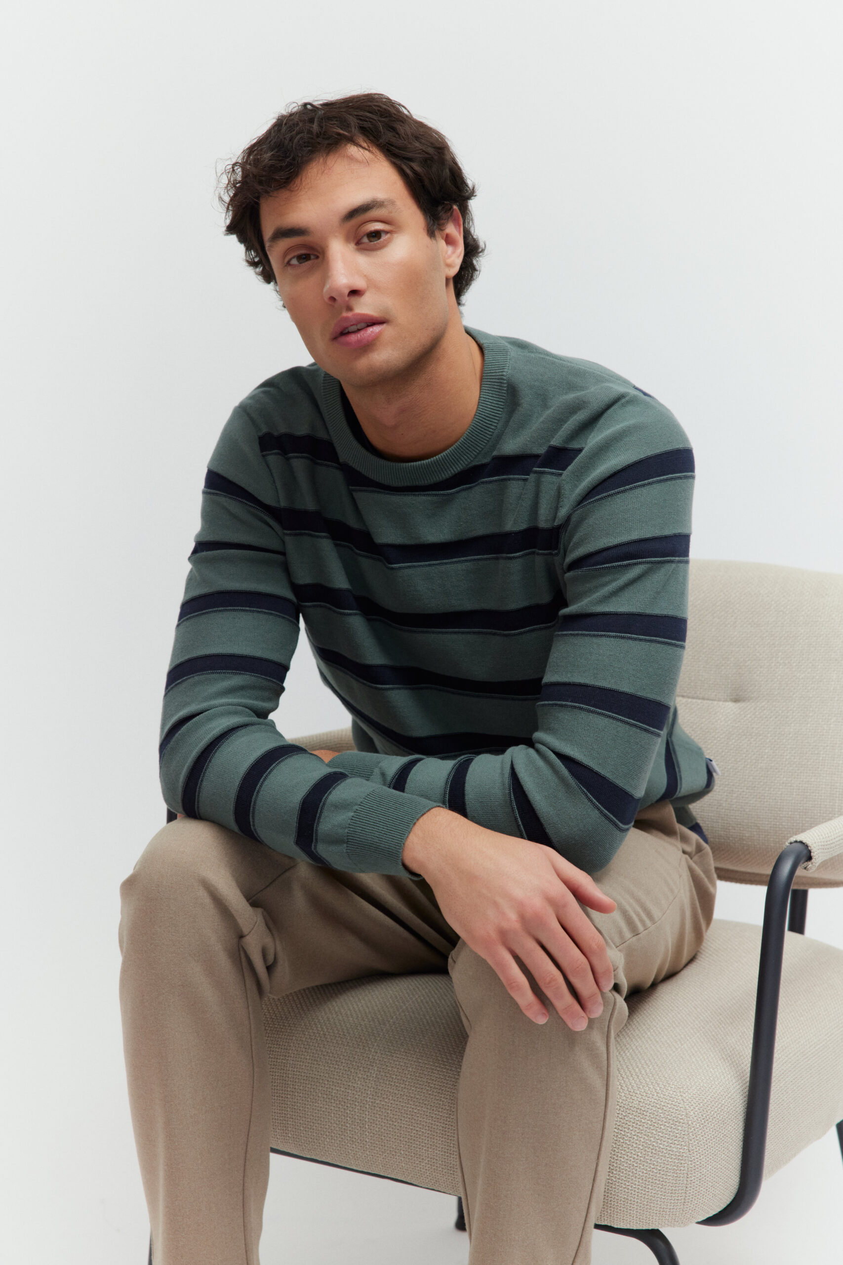 Model sitzt auf einem hellen Sessel in einem grün-blau gestreiften Pullover und hellen Hosen.