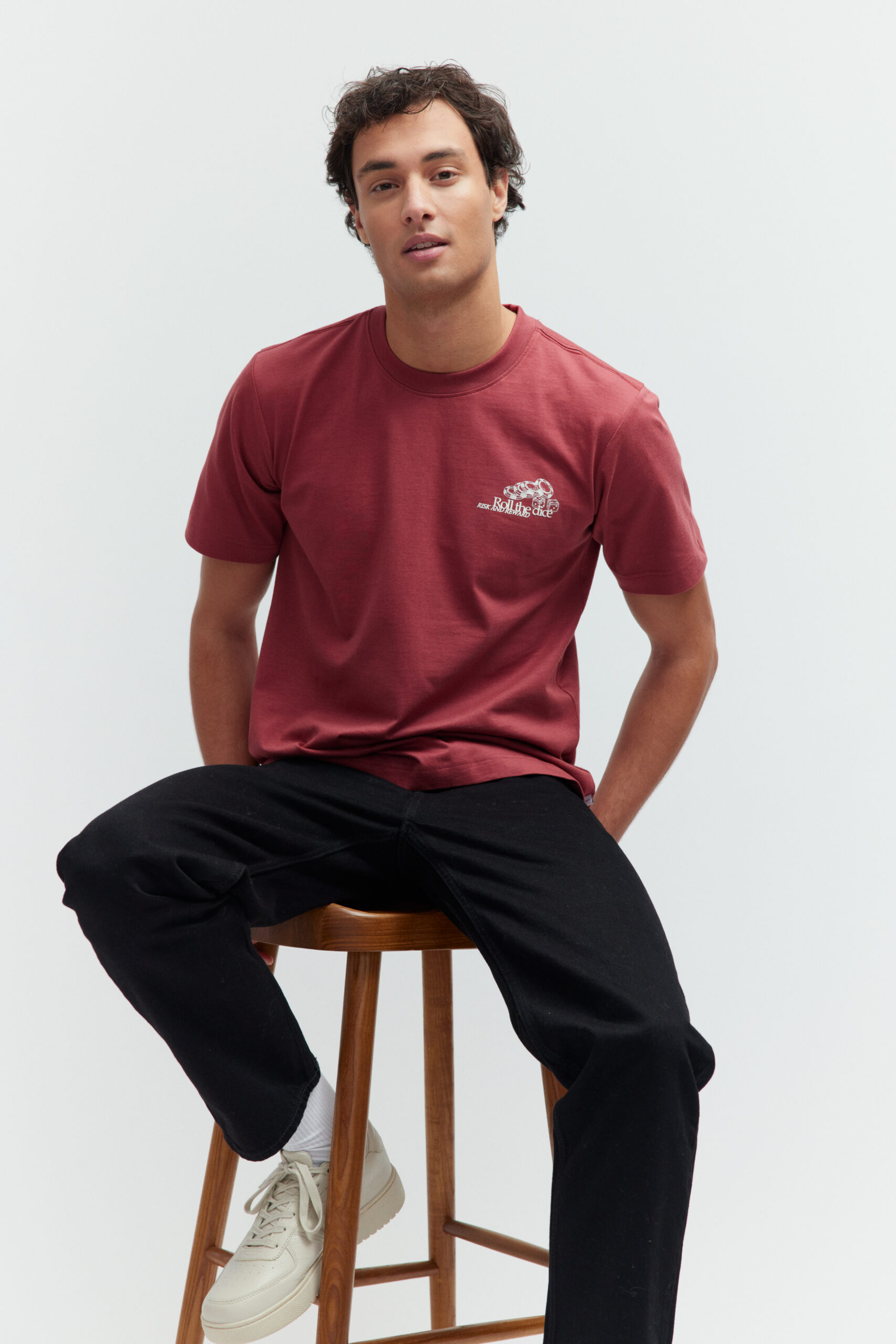 Model sitzt auf einem Holzschemel in bordeauxrotem T-Shirt und schwarzer Jeans.