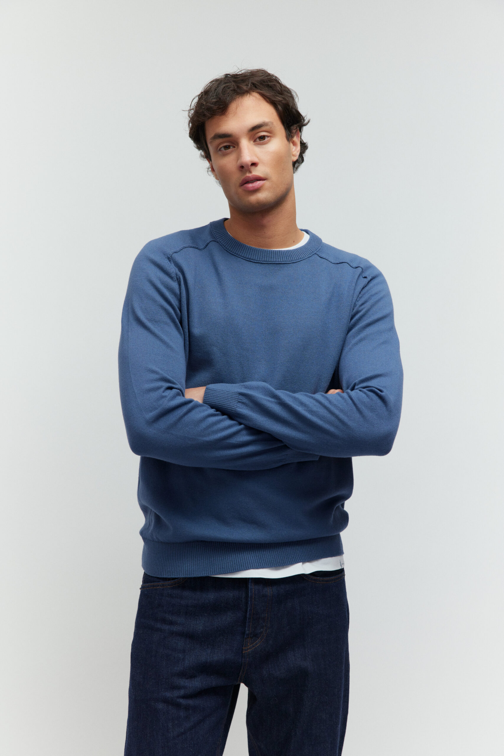 Model in blauem Pullover und dunkler Jeans, die Arme vor der Brust verschränkt.