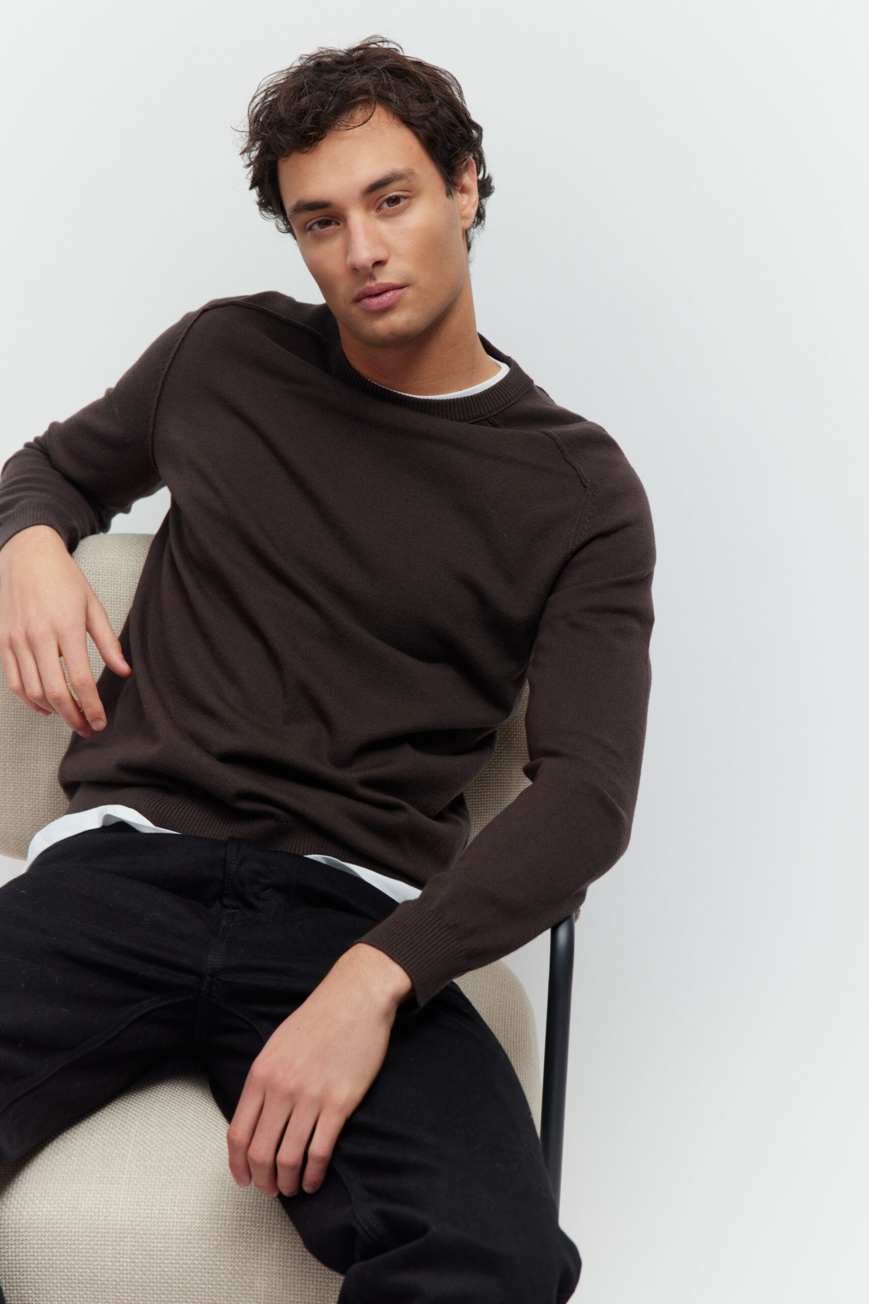 Model sitzt auf einem hellen Sessel in moccabraunem Pullover und schwarzer Jeans.