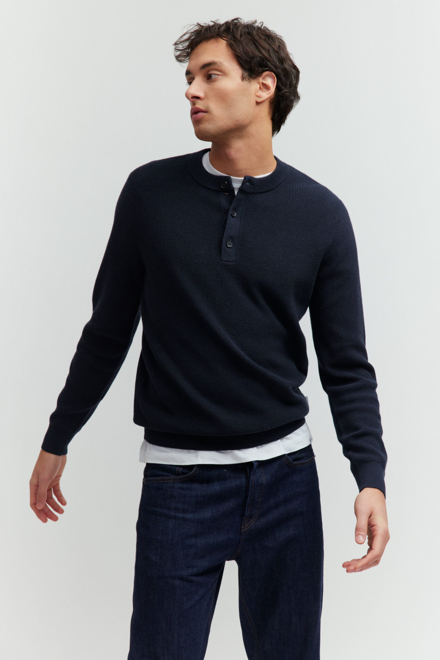 Model in dunkelblauem Henley-Pullover mit Knöpfen und dunkler Jeans vor hellem Hintergrund.